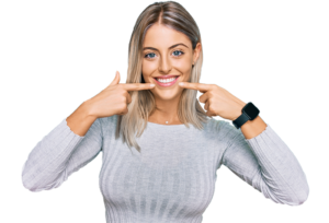 A smiling woman in a gray sweater points to her mouth. She wears a smartwatch, standing against a black background. Tone is happy and inviting.